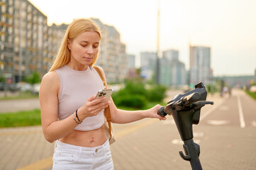 woman using smartphone while holding electric scooter in urban setting. casual fashion meets mobility, showcasing modern city transportation and tech lifestyle on a sunny day
