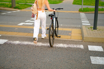 woman with bicycle crossing urban street. young female in white pants, tan bag, pushes bicycle over...