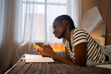 Young african-american woman is lying on her bed, using her smartphone with an open notebook next...