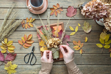 Top view of hands folding dried leaves and flowers into rustic autumn bouquet on wooden table with foliage, scissors and cup of coffee. Cozy autumn atmosphere. Perfect for crafts or seasonal content