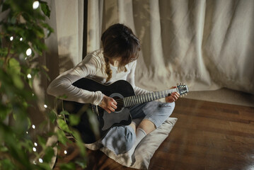 Young woman with braided hair playing a black acoustic guitar sitting on the floor by the curtains...