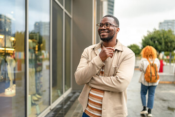 Smiling black man looks at store window while strolling in modern urban area during overcast...
