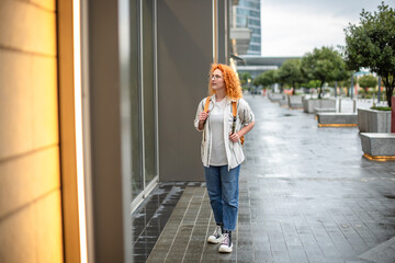 Ginger woman with orange hair explores urban street on a rainy day, admiring storefronts in a...