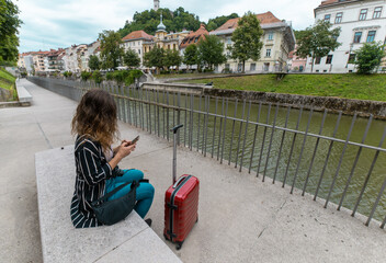 Young woman with red suitcase sitting near a calm river in Slovenia, checking her phone, surrounded...