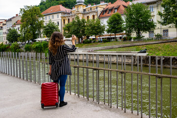Female traveler with red suitcase taking photos of traditional buildings along Ljubljana riverbank,...