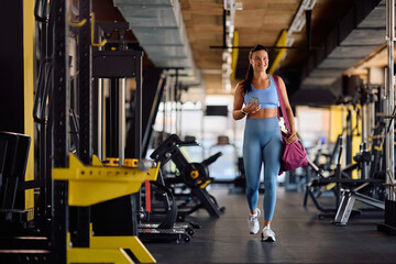 Happy sportswoman with mobile phone and sports bag walking through health club.
