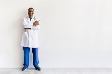 Full body portrait of smiling African American doctor pointing aside at copy space against white background. Confident professional promoting service or directing attention