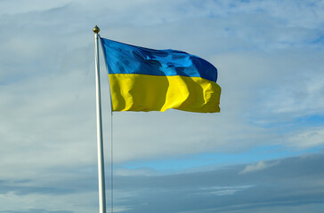 A vibrant Ukrainian flag displays its blue and yellow colors boldly as it waves in the breeze. The backdrop features a mix of clouds and blue sky, creating a serene atmosphere