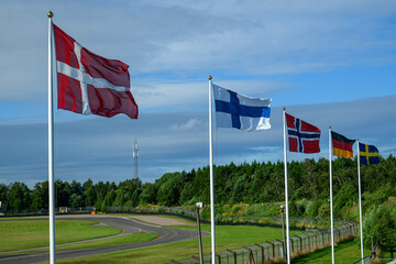 Various flags representing Nordic countries flutter in the wind alongside a racetrack surrounded by...