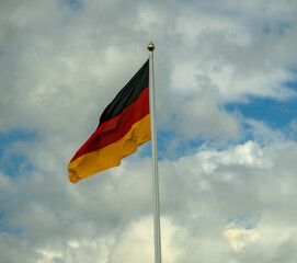 The vibrant German flag is prominently displayed on a flagpole, fluttering gracefully against a backdrop of fluffy clouds and blue skies, illustrating national pride on a sunny afternoon