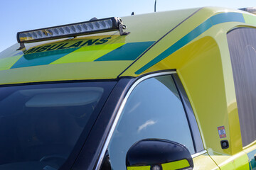 An emergency ambulance vehicle is parked at the roadside under a clear blue sky. The bright yellow and green design is prominent, illustrating readiness for medical emergencies