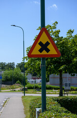 A caution sign placed on a street pole alerts pedestrians and drivers to intersection ahead. The setting features lush green trees and a well-maintained sidewalk