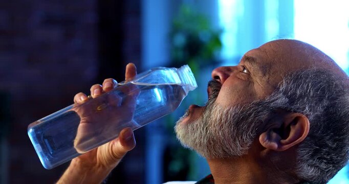 Indian senior man drinking water from glass bottle while sitting at table in modern home, happy and healthy aged asian male staying hydrated indoors during peaceful relaxed lifestyle in retirement