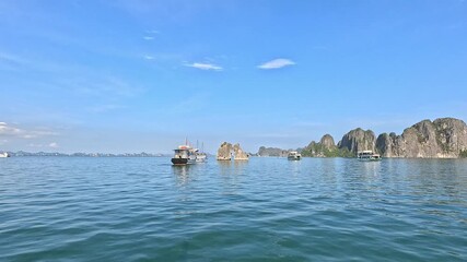 Panning shot of tourist boats and stunning limestone islets, including the iconic Kissing Rocks, in Ha Long Bay, Vietnam, under a clear blue sky.