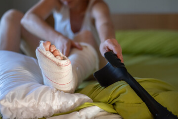 Woman sitting in bed gently touching her broken leg and grabbing a crutch, getting ready to deal...