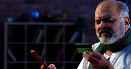 Indian senior man using credit card and smartphone for secure online transaction while sitting at table in modern home, happy aged asian male enjoying digital banking and internet shopping - Powered by Adobe
