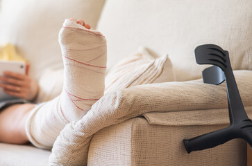 Patient is recovering from a broken leg injury at home, resting on a sofa with a cast and crutches nearby, using a smartphone for entertainment or communication