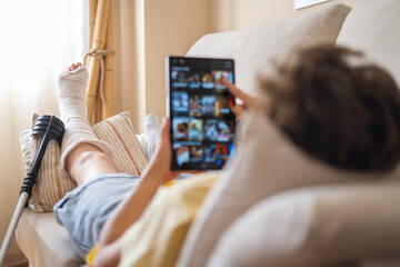 Young patient is lying on sofa with broken leg in cast, using digital tablet and browsing online...
