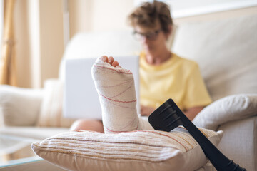 Senior woman resting on a sofa at home, managing recovery from a broken leg in a cast. Using a...