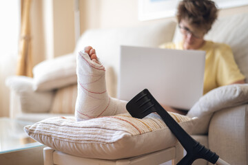 Woman resting with broken leg supported on cushion, using crutches and working from home using...