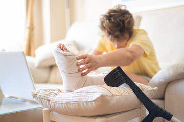 Woman sitting on the sofa, gently touching her broken leg encased in a plaster cast, with crutches...