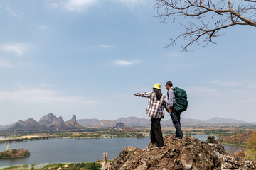 rear view of a traveling couple holding hands on a rocky summit, gazing at the landscape below that...