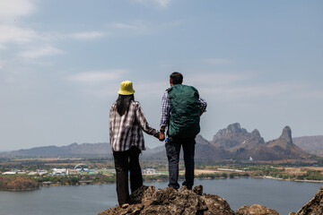 rear view of a traveling couple holding hands on a rocky summit, gazing at the landscape below that...