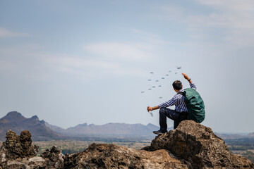 male traveler with a backpack is sitting on a rocky peak, raising a hand as if waving or greeting...