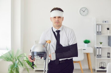 Portrait of injured young man standing in office with his arm in plaster cast and bandaged head...