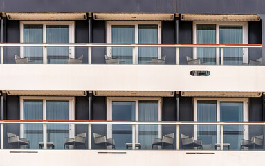 side of a cruise ship with balconies and deck chairs. Cabin windows with tables and seats on the...