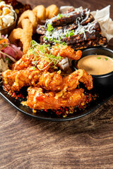 Close-up of crispy fried chicken wings with sauce, onion rings, and herbs served on a black plate over rustic wooden table, styled as gourmet comfort food.