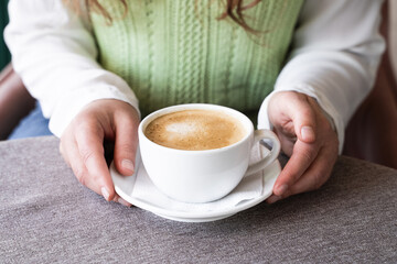 Woman's hands holding a white cup of cappuccino with creamy coffee foam on a saucer in a cozy cafe. Coffee break, love for caffeine, pleasure of enjoying a warm beverage in a relaxing atmosphere.