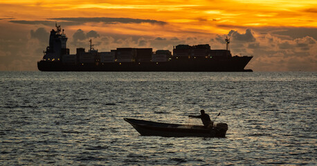 A lone fisherman casts his net at dawn while a large cargo ship passes in the distance,...