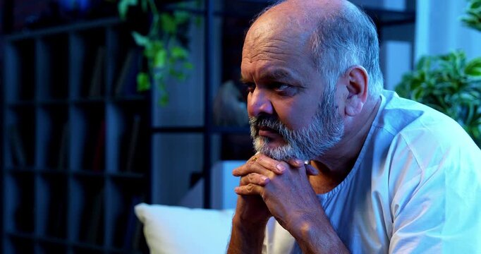 Senior Indian man deep thinking while sitting on sofa in bright airy modern home, thoughtful Asian elderly person with worried facial expression, old retired man lost in serious thoughts indoors