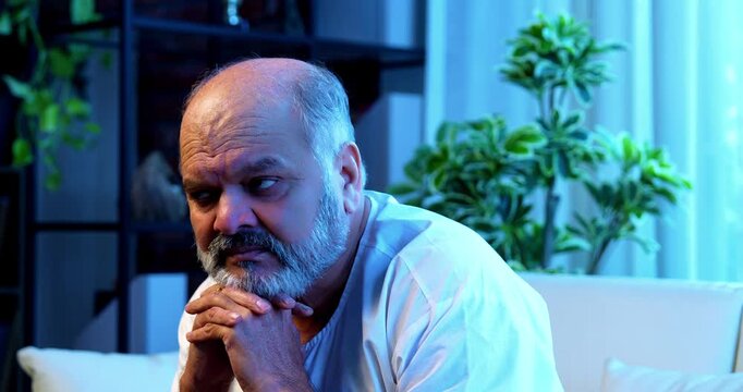 Senior Indian man deep thinking while sitting on sofa in bright airy modern home, thoughtful Asian elderly person with worried facial expression, old retired man lost in serious thoughts indoors