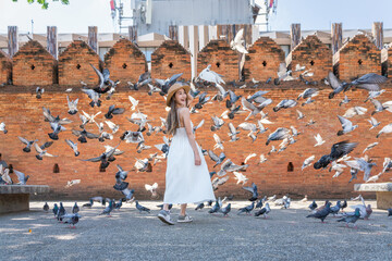 Asian tourist woman enjoying travel moments at Tha Phae Gate, Chiang Mai. Young woman in white dress surrounded by flying pigeons in historical city square.