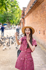Young Asian woman video calling with phone at Tha Phae Gate in Chiang Mai. Female tourist waving...