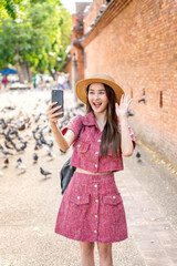 Young Asian woman video calling with phone at Tha Phae Gate in Chiang Mai. Female tourist waving...