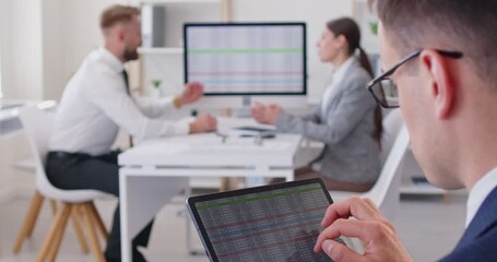 Close-up shot of male analyst wearing glasses looking through the information on spreadsheets on his tablet, while his colleagues disscuss indicators and data on a presentation monitor with statistics - Powered by Adobe