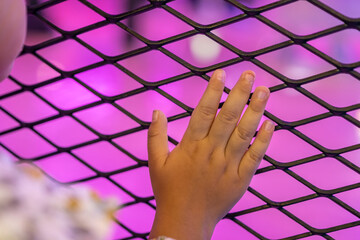 Close-up of a child's hand touching a black mesh fence with a vivid pink and purple neon...