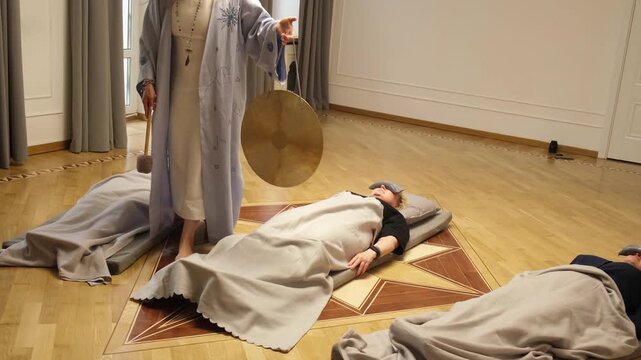 Three people lying peacefully on mats during a gong meditation session. A person stands nearby with a gong and mallet, creating a calming and serene environment for relaxation