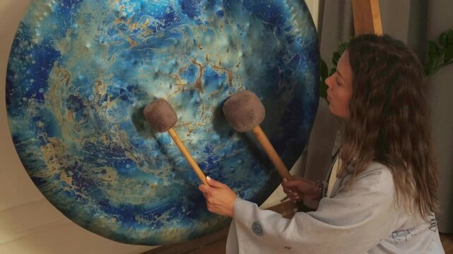 Woman using mallets to play a large gong, creating soothing sounds for a sound bath session. The focus is on the gong's vibrant colors and circular patterns