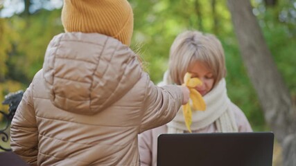 close up of woman sitting on bench working on laptop outdoors in autumn park while daughter offers yellow leaves to distract mother who gently shifts hand aside before child withdraws - Powered by Adobe