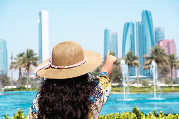A woman in a straw hat and floral dress points at skyscrapers near a blue pool, with palm trees...