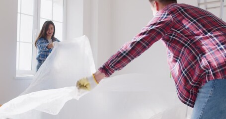 Young family couple covering the floor with white plastic protection film before they start painting walls in the room. House renovations, DIY, decoration, home improvement concept - Powered by Adobe
