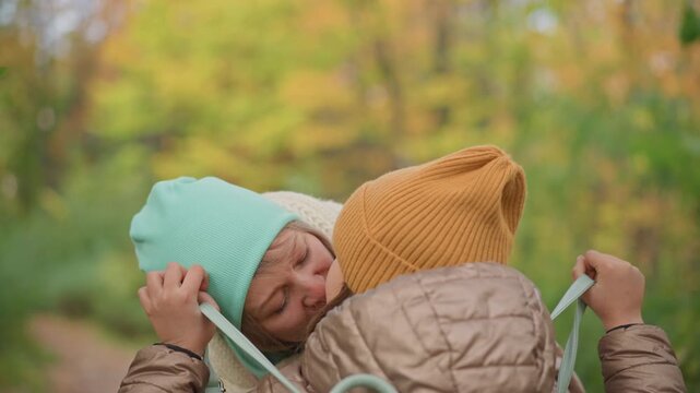 tender moment as mother leans in to kiss daughter on forehead while both wear cozy winter hats and jackets, surrounded by soft-focus fall foliage in peaceful forest setting