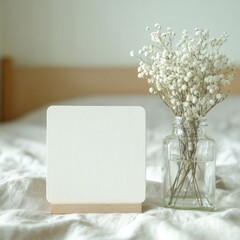 Blank square card with wooden stand sits on linen fabric beside small glass vase of white baby's breath flowers