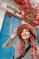 Young woman tourist wearing hat in front of a traditional blue door and red flowers during summer vacation