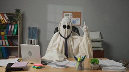 Halloween businessman ghost in white sheet sits at office desk looks at camera holding hand up and pointing finger. Happy halloween business concept.