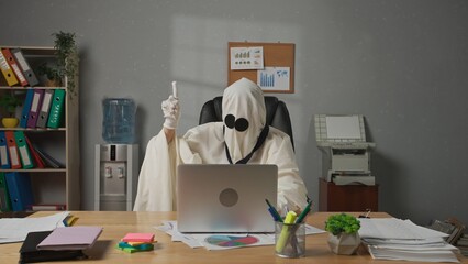 Halloween businessman ghost in white sheet sits at office desk looks at camera holding hand up and...
